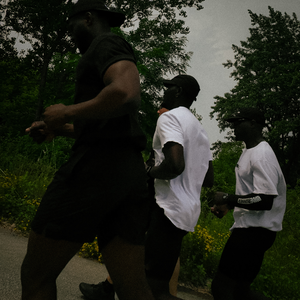 Three men jogging outdoors on a paved path surrounded by green trees under an overcast sky. Two men wear black athletic wear, and one wears a white t-shirt and black shorts.